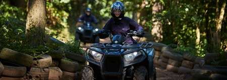 ATV rider drives along a narrow, tire-lined dirt track, navigating through a shaded forest; another rider follows behind among trees, sunlight filtering through leaves.