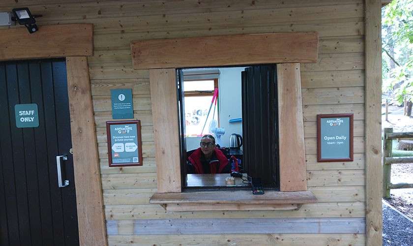 Ticket attendant sits behind a wooden kiosk window, ready to assist; surrounding signs read: “STAFF ONLY” and “Open Daily,” with additional smaller, illegible text on two framed notices.