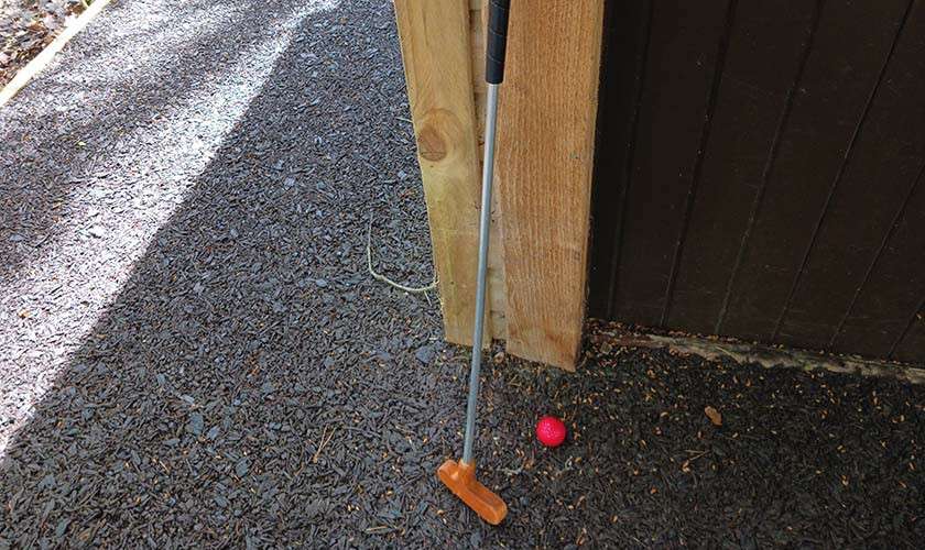 Mini-golf putter leans against a wooden post, resting upright. Nearby, a bright pink ball sits on dark gravel beside a wooden wall along a shaded outdoor pathway.