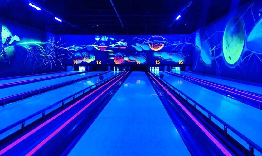 Bowling ball rolls down a neon-lit lane; pins stand ahead; cosmic murals glow under blacklight; lane numbers read 12, 13, 14, 15, 16.