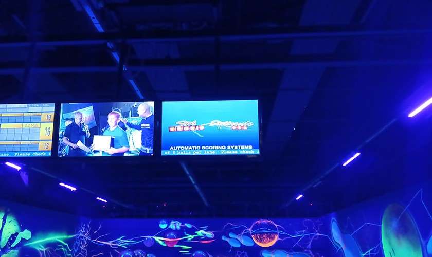 Overhead monitors display bowling details; one shows scores, another a video feed, another a blue screen reading “AUTOMATIC SCORING SYSTEMS.” Visible numbers: 19, 16, 12; text: “Please check.” Neon blacklight alley.