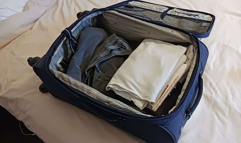 Open navy suitcase holds folded clothes—white T-shirts and denim—arranged neatly; mesh pockets in the lid are visible; situated on a bed in a simple bedroom.
