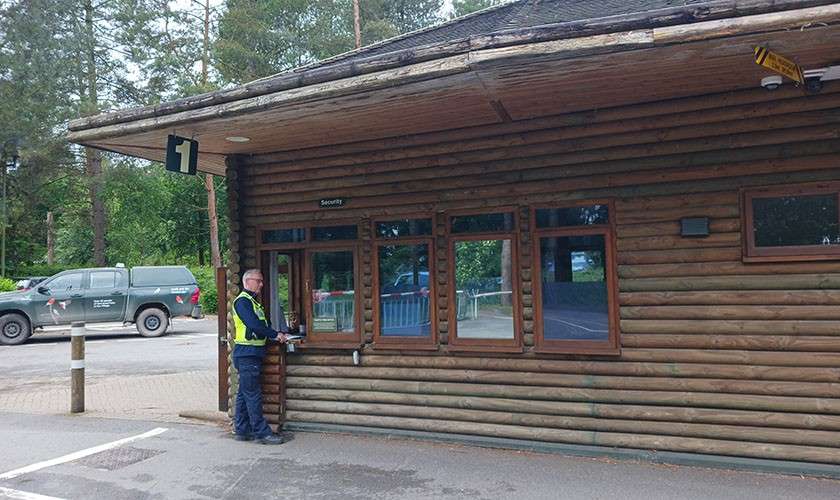 Security officer unlocks a windowed log-cabin booth. Surroundings show a parking area with a green pickup and trees. Visible text: "Security", "1", "Center Parcs".