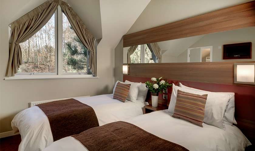 Twin beds sit side-by-side, dressed with white linens and brown throws; bedside flowers and wall sconces accent them, in a sloped-ceiling room with large curtained window and forest view.