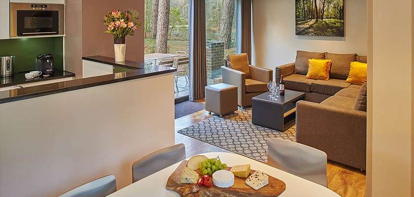 Open-plan living room displays an L-shaped sofa with yellow cushions, facing a coffee table with wine. In context, adjoining kitchen and dining area, sliding doors leading to a forested patio.