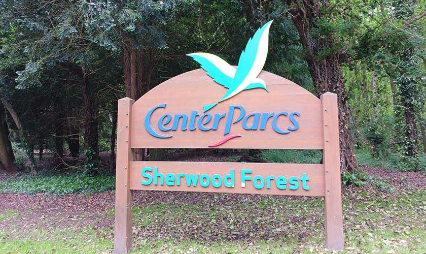 Wooden entrance sign displays “Center Parcs” and “Sherwood Forest,” standing among tall trees and leaf-strewn ground at a forest resort entrance.