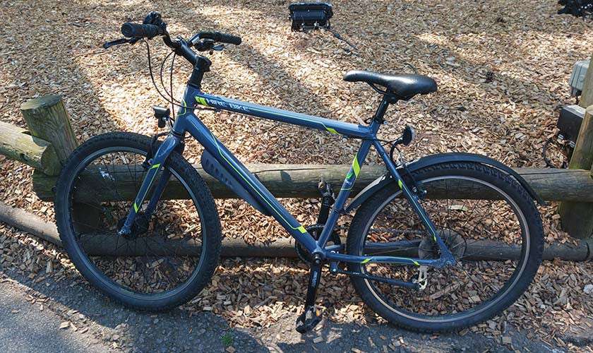 Mountain bike leans against a wooden fence, parked on a paved path beside wood-chip ground; front suspension fork, flat handlebars, knobby tires, disc brakes, and kickstand are visible. No legible text visible.
