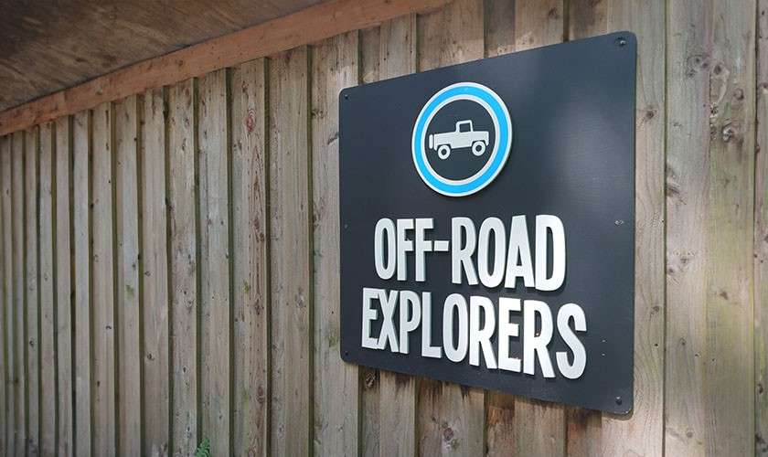 Sign displays "OFF-ROAD EXPLORERS" with a pickup-truck icon, mounted on a vertical wooden fence under an overhang, catching daylight.