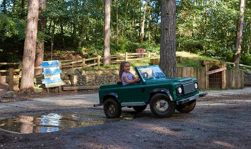 Child driving a small green toy off-road jeep, smiling while steering. The vehicle splashes near a puddle on a gravel path within a wooded park, fences and trees surrounding.