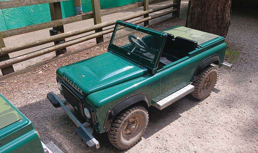 Small green ride-on jeep sits parked, steering wheel visible, windshield upright; mud-streaked tires rest on gravel. Wooden fence and tree surround. Visible text: “DEFENDER” on the front.