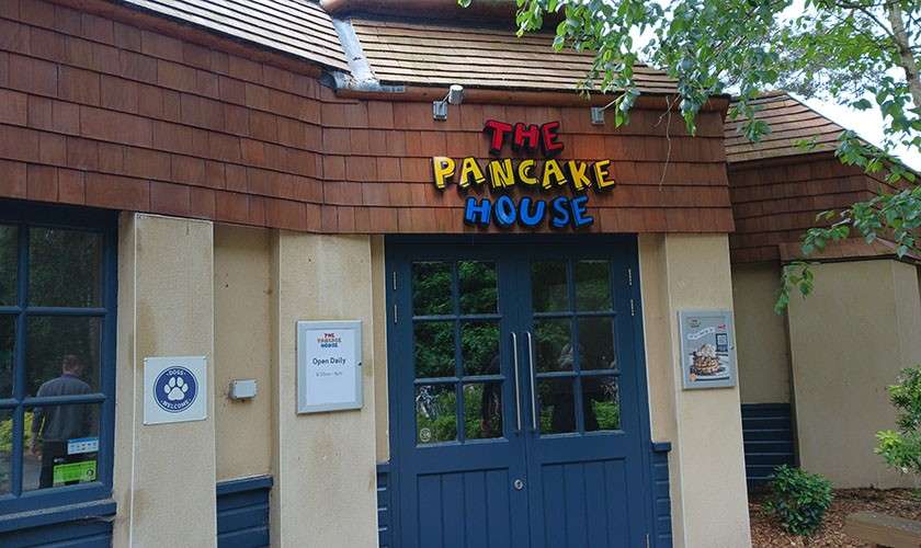Restaurant entrance displays colorful “THE PANCAKE HOUSE” sign; closed double doors. Wall signs: “Dogs Welcome”; “Open Daily” (other text illegible); right poster with pancake image (text unreadable); leafy surroundings.