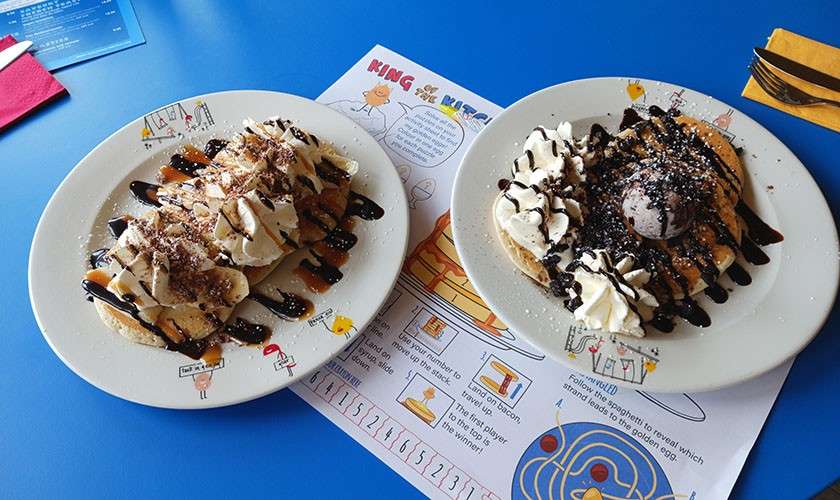 Two dessert waffles sit on plates—drizzled with chocolate and caramel, topped with bananas, whipped cream, and ice cream—on a blue table over a children’s activity placemat with utensils.

Text: “KING OF THE [illegible]”; “1 2 3 4 5 6 7 8 9 10 11 12 13 14 15 16”. Other small text is too unclear to read.