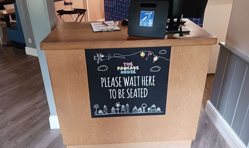 Reception counter displays a sign: "THE PANCAKE HOUSE PLEASE WAIT HERE TO BE SEATED"; tablet and clipboard rest on top, with dining area and blue-tiled wall in the background.
