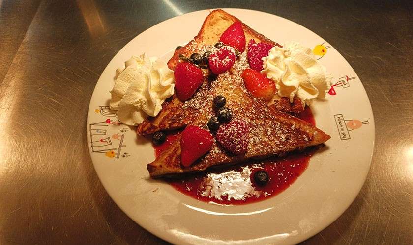 French toast slices top with strawberries, blueberries, and powdered sugar, resting in berry sauce, flanked by whipped cream. On a white plate with small illustrations. Text: “DAY”; “happy day”.