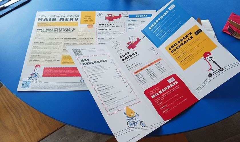 Colorful menus lie open, displaying choices, on a round blue table with cutlery and a pink napkin nearby.

Text visible:
- THE PANCAKE HOUSE
- MAIN MENU
- AMERICAN STYLE PANCAKES
- BELGIAN STYLE WAFFLE
- EXTRAS
- DUTCH STYLE SAVOURY PANCAKE
- OMELETTES
- HOT BEVERAGES
- SOFT DRINKS
- SMOOTHIES
- CHILDREN’S COCKTAILS
- ICED MILKSHAKES
