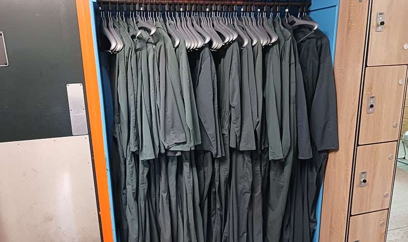 Coveralls hang uniformly on silver hangers, tightly grouped on a rail inside a blue locker alcove, with a metal door on the left and wood lockers on the right.
