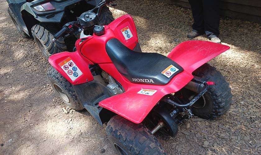Red all-terrain vehicle sits parked; seat labeled HONDA; warning decals visible; located on a dirt surface beside another ATV and a wooden structure.