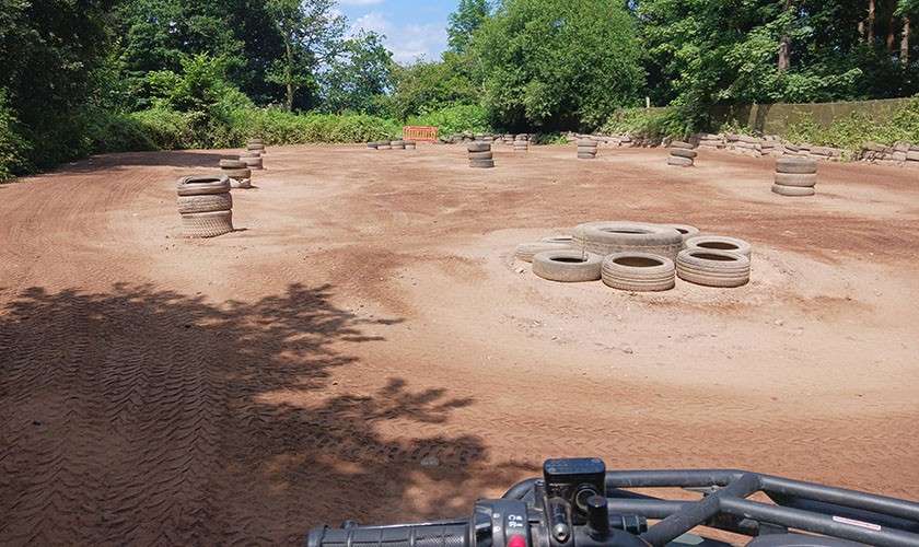 ATV handlebars point forward, navigating a dusty track; stacked tires mark turns within a clearing bordered by trees and shadows under a sunny sky.