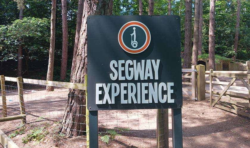 Sign displays "SEGWAY EXPERIENCE," standing by a forest trail, beside wooden fences and gates among tall trees.