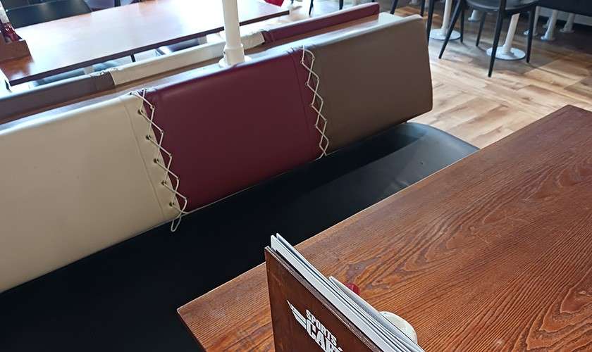 Menu booklet stands upright on a wooden table; behind it, a laced cream-and-maroon booth bench sits in a restaurant with wood flooring. Visible text on menu: "ROAD CAF..."