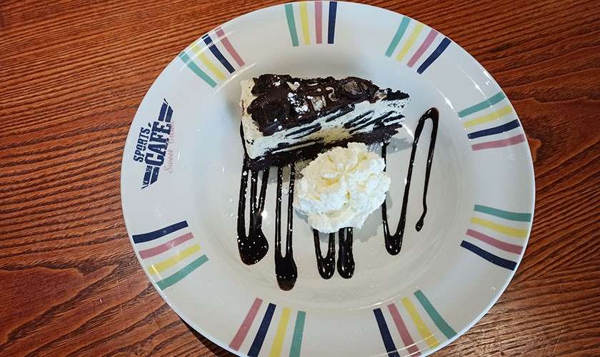 Chocolate-drizzled cookies-and-cream cheesecake slice rests beside a dollop of whipped cream, on a white plate with colorful stripes, text: “SPORTS CAFE,” set on a wood-grain table.