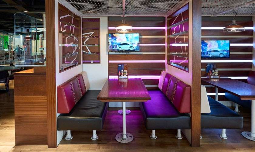 Booth seats flank a wooden table facing wall-mounted TVs. Pink LED strips glow. In a modern diner, pendant lamps hang overhead, condiments on the table, and wood partitions frame booths.