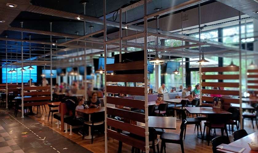 Diners sit and talk at tables, separated by wooden slat partitions; pendant lamps hang above in a spacious, modern restaurant with large windows, TVs, and a patterned floor walkway.