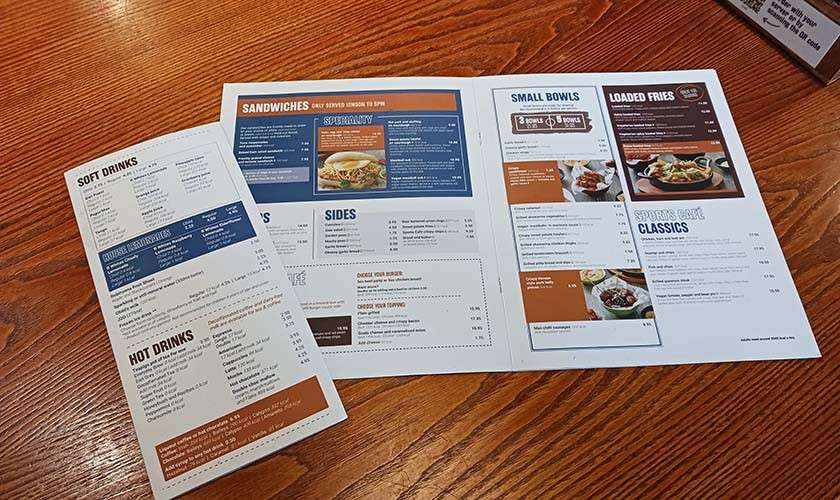 Menus lie open, showing printed categories on a wooden table. Text: SOFT DRINKS, HOT DRINKS, SANDWICHES, SIDES, SMALL BOWLS, LOADED FRIES, CLASSICS. Additional smaller text is present but unreadable.