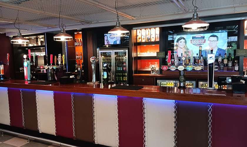 Beer taps line a long wooden bar, standing idle; behind them, fridges and shelves of bottles sit under hanging lights and a television displaying text: “SCOOP.”