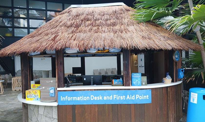 Thatched-roof service hut provides assistance, staffed counters with computers in front; context: tropical-themed lobby with palms and stone flooring. Visible text: Information Desk and First Aid Point.