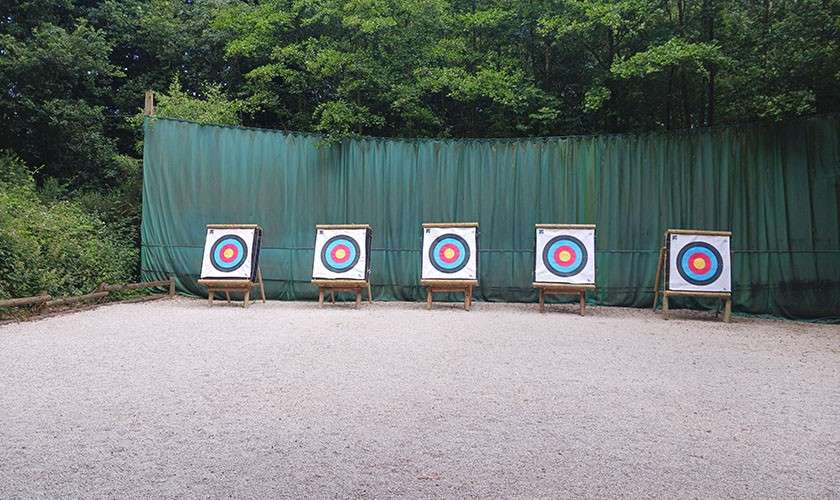 Five colorful archery targets stand on wooden easels, aligned side by side, facing the shooter; a green safety curtain and surrounding trees enclose the gravel practice range.