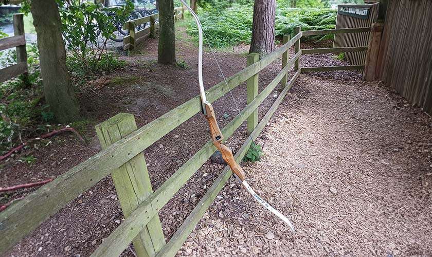 A recurve bow hangs by its string over a wooden fence, angled downward; context: tree-lined outdoor path with wood-chip ground, railings, and a nearby enclosure in a forested area.