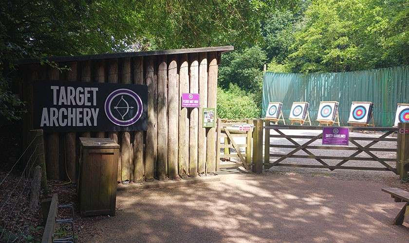 Targets stand aligned for practice in a fenced outdoor archery range beside a log-sided shed and gate. Text: TARGET ARCHERY; SAFETY GUIDELINES.