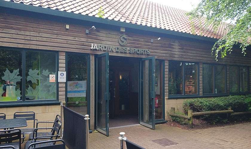 Glass double doors stand open, inviting entry; a wooden-clad building houses facilities, surrounded by trees and patio seating. Text: JARDIN DES SPORTS.