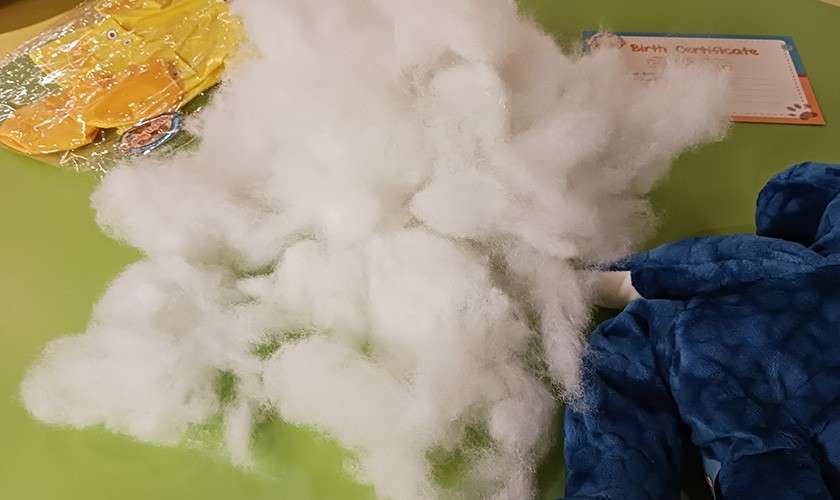 Polyfill stuffing sprawls across a green table, beside an unstuffed blue plush body and a packaged yellow outfit; a card reads: "Birth Certificate".