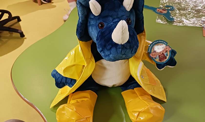 Blue plush triceratops sits upright, wearing a yellow raincoat and matching rain boots, on a green table in a playroom; craft materials and a chair are visible around it.