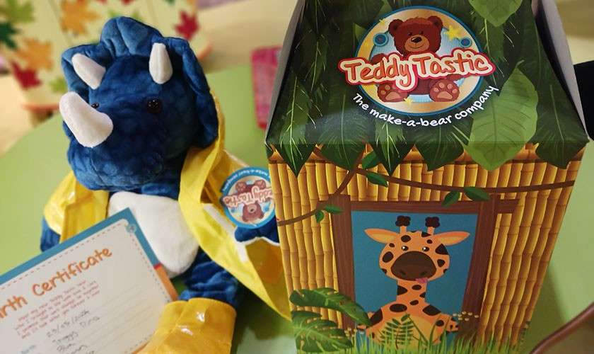 Plush blue triceratops sits wearing a yellow raincoat beside a jungle-themed box and certificate on a table. Visible text: “TeddyTastic,” “The make-a-bear company,” “Birth Certificate.”