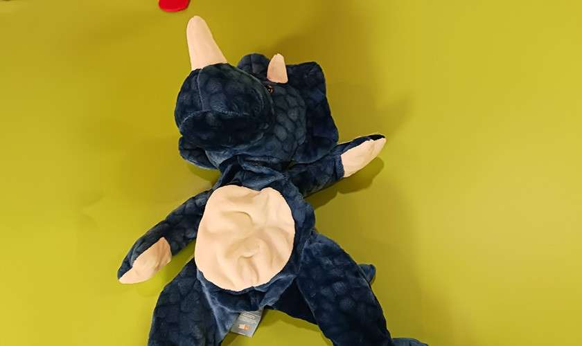Blue triceratops plush lies belly-up, limbs splayed, on a bright green surface; beige horns, belly, and feet visible. A small red item peeks near the top edge.