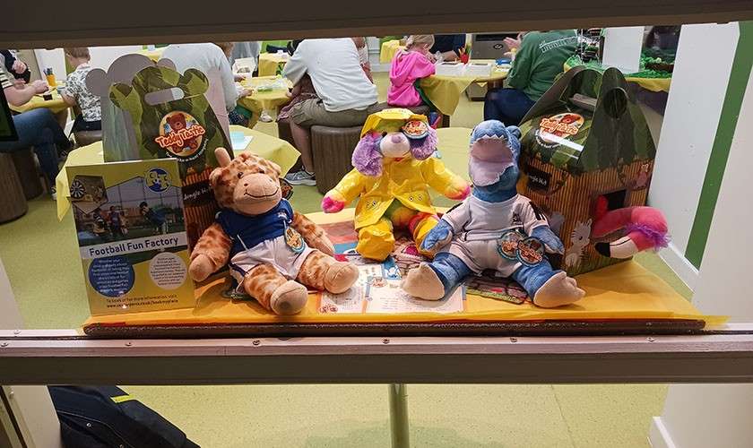 Plush giraffe, dog, and dinosaur sit on a promotional display; people gather at tables behind. Text: TeddyTastic; Football Fun Factory.