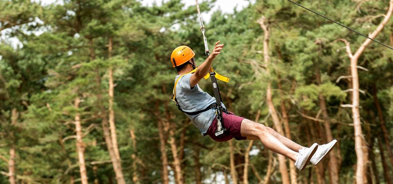Zip-liner glides on a cable, wearing a harness and orange helmet labeled “CAMP,” above a dense forest of tall pine trees in daylight.