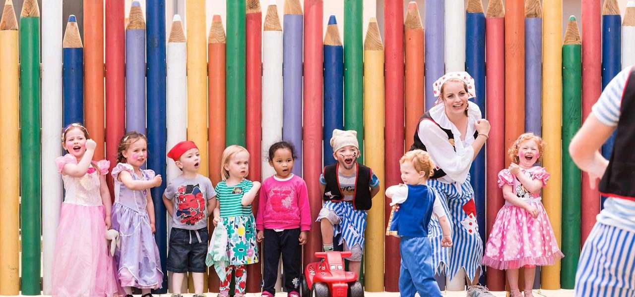Children and a costumed adult laugh and gesture while standing in a row, some in princess or pirate outfits, before a fence of colorful giant pencils at an outdoor celebration.