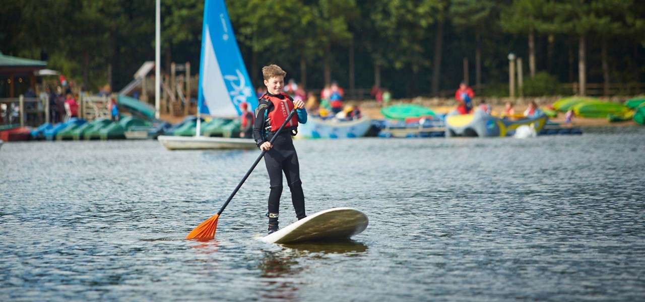 Child on a stand-up paddleboard paddles, wearing a life vest, on a calm lake; behind, people use kayaks and a sailboat with blue sail reading Pico near a wooded shore.