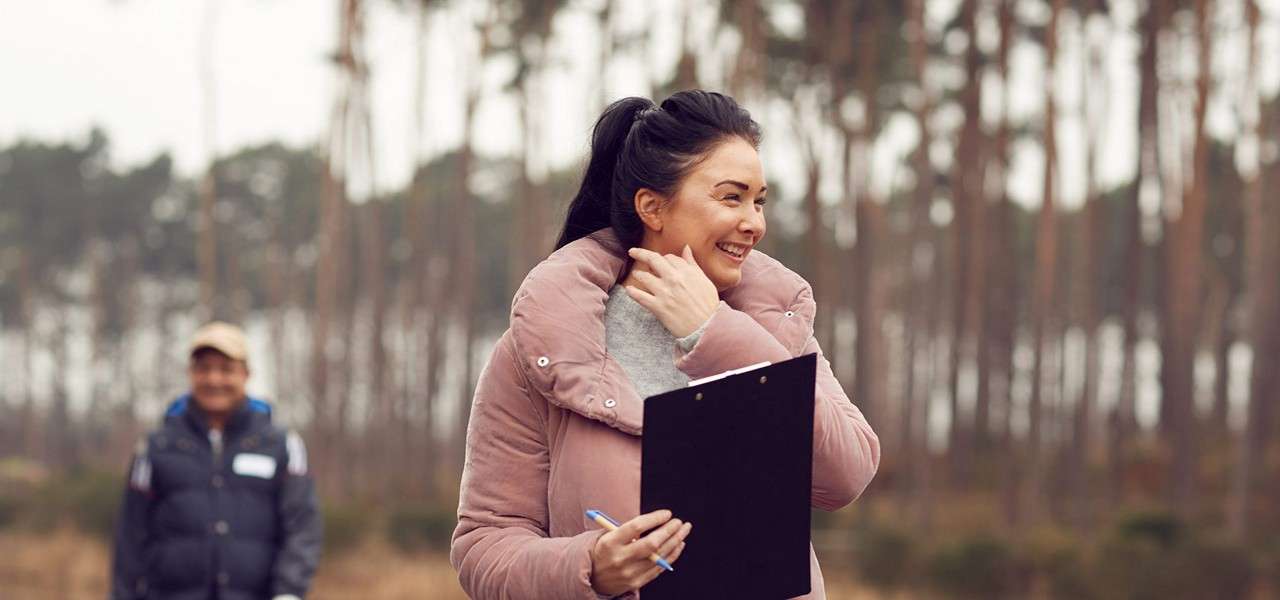 Person in pink puffer jacket smiles while holding a clipboard and pen, hand near face; outdoors in a pine forest with another bundled person blurred in the background.