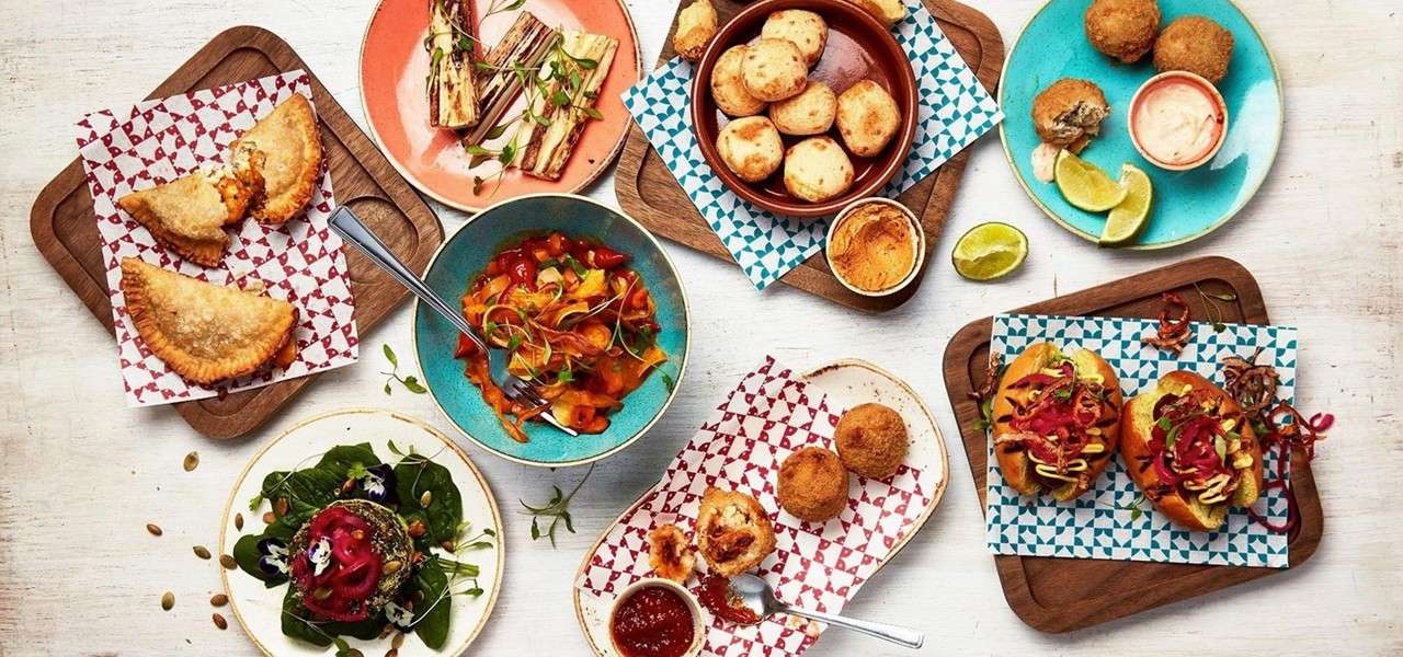 Assorted small dishes arranged on a tabletop: empanadas, sliders, arancini, soup, salad, grilled vegetables, and cheese breads; served on colorful plates with dipping sauces, lime wedges, and wooden boards.
