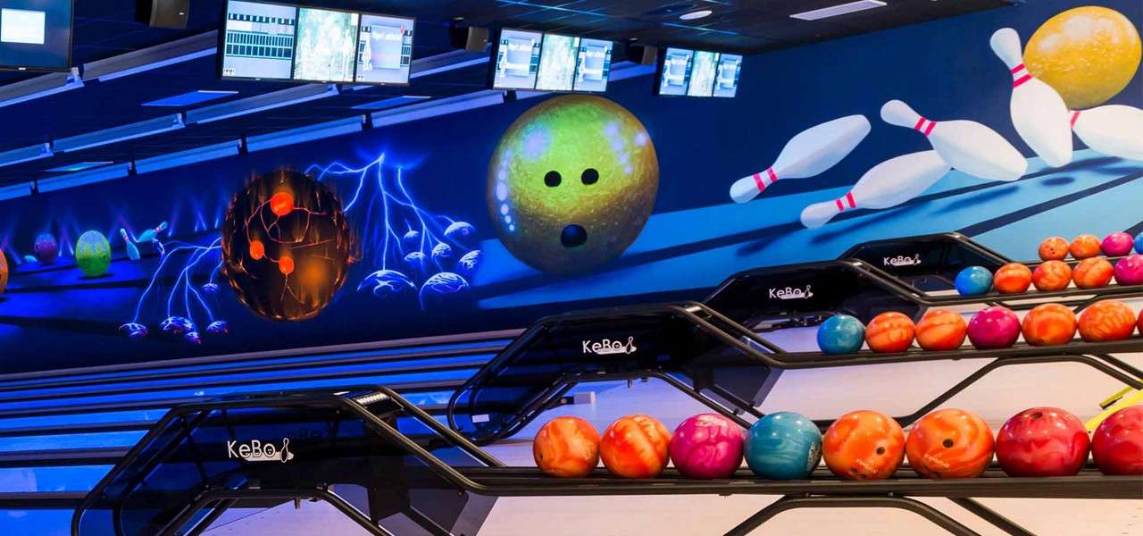Bowling balls rest on black racks labeled “KeBo,” while pins and balls animate across a vibrant wall mural; overhead screens hang above multiple lanes in a neon-lit bowling alley.