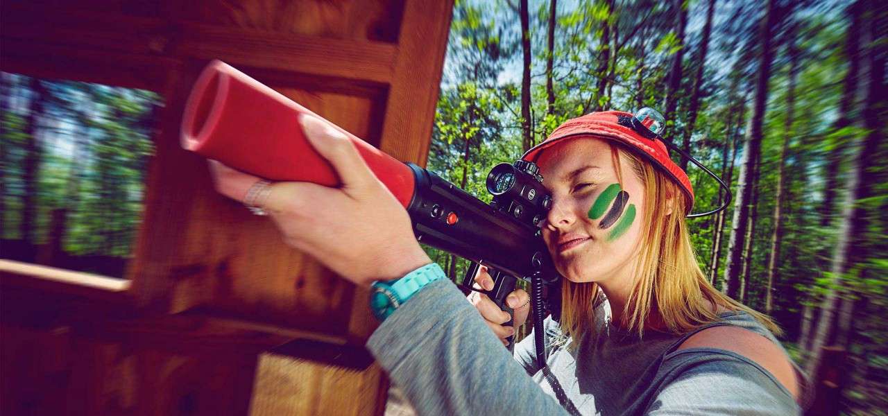 Person with face paint aims a laser tag rifle through a wooden bunker opening, squinting through a scope. Surrounding forest blurs with motion, indicating action in an outdoor game arena.
