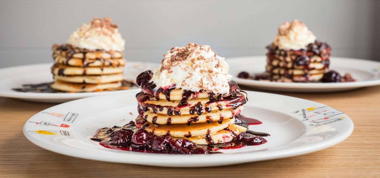 Pancake stack drips berry sauce and chocolate, topped with whipped cream and shavings, sitting on a white plate; two similar stacks blur in the background.