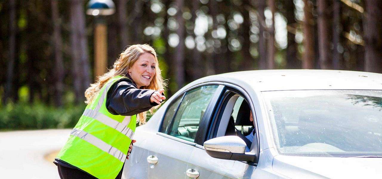 Safety-vested attendant leans toward a silver sedan, smiling and pointing to direct the driver, along a tree-lined road or parking area in daylight.