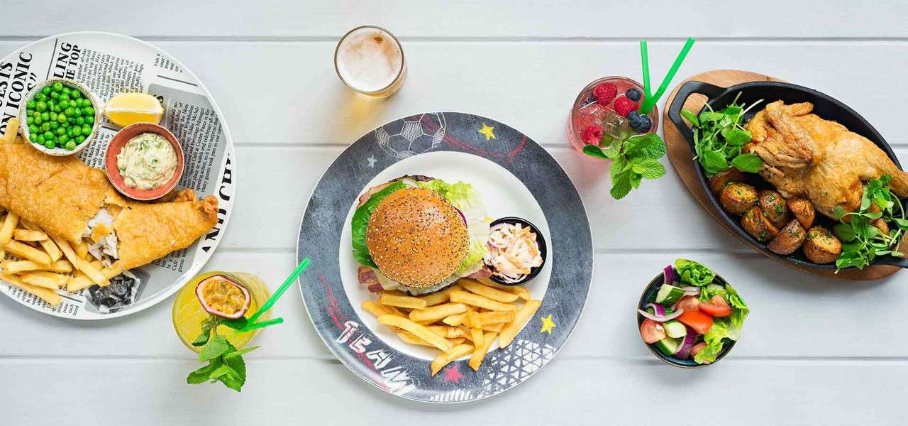 Burger with fries and coleslaw sits center; fish and chips with peas left; roast chicken with potatoes right; salads and fruity drinks surround on a white table. Text: TEAM.