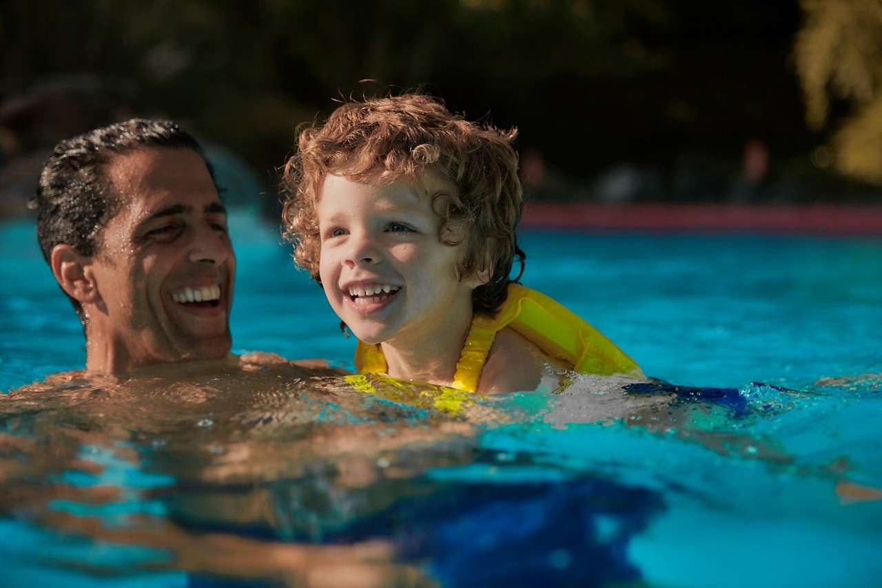 Child in a yellow flotation vest smiles while floating and splashing, supported by an adult. They enjoy a sunlit outdoor swimming pool with blue water and blurred greenery.